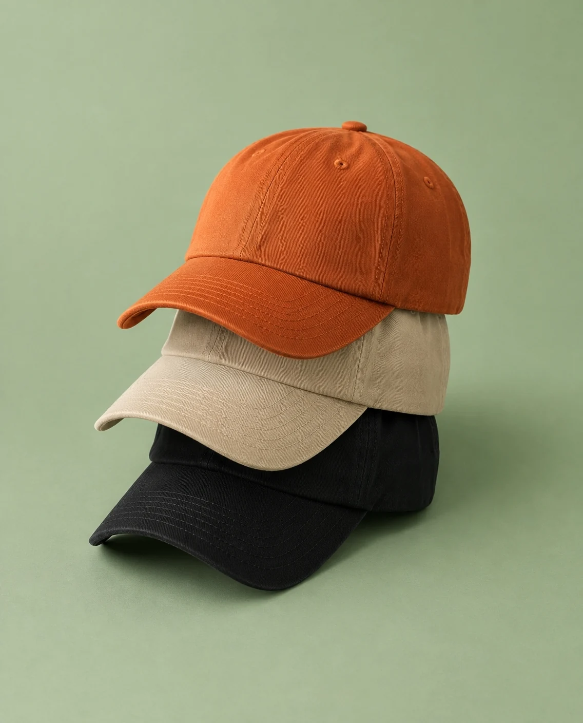 Tres gorras lisas apiladas en naranja, arena y negro sobre fondo verde salvia